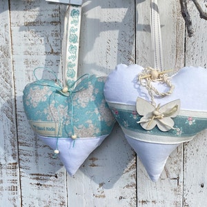 Könnte beinhalten: Zwei handgefertigte Stoff-Anhänger in Herzform mit einem blauen und weißen Blumenmuster. Ein Herz hat einen weißen Schmetterling und das andere ein Schild mit der Aufschrift "Hand Made".
