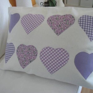 Könnte beinhalten: Ein weißes Kissen mit acht aufgenähten lila und weißen Herzen. Die Herzen haben verschiedene Muster, darunter Gingham, Blumen und einfarbig.
