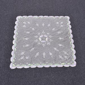 Può includere: Un centrino quadrato, trasparente, bianco, con bordo smerlato. Il centrino presenta un delicato motivo floreale e a foglie con un disegno centrale a stella. I bordi hanno un sottile bordo verde e bianco.