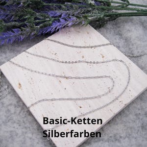 Puede incluir: Collares plateados con diferentes estilos de cadena se muestran sobre una superficie cuadrada y clara. Los collares están colocados, con una ramita de flores moradas en el fondo. El texto "Basic-Ketten Silberfarben" es visible.