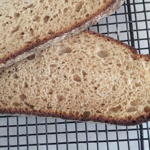May include: Two slices of freshly baked bread with a golden-brown crust and a porous interior. The bread rests on a black wire cooling rack, showcasing its texture and freshness. The crust is a darker shade of brown, indicating a perfect bake.