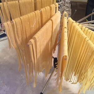 May include: Freshly made pasta noodles hanging on a drying rack. The noodles are a light yellow color and are arranged in neat rows. The rack is made of metal and plastic, and the background is a light-colored surface.