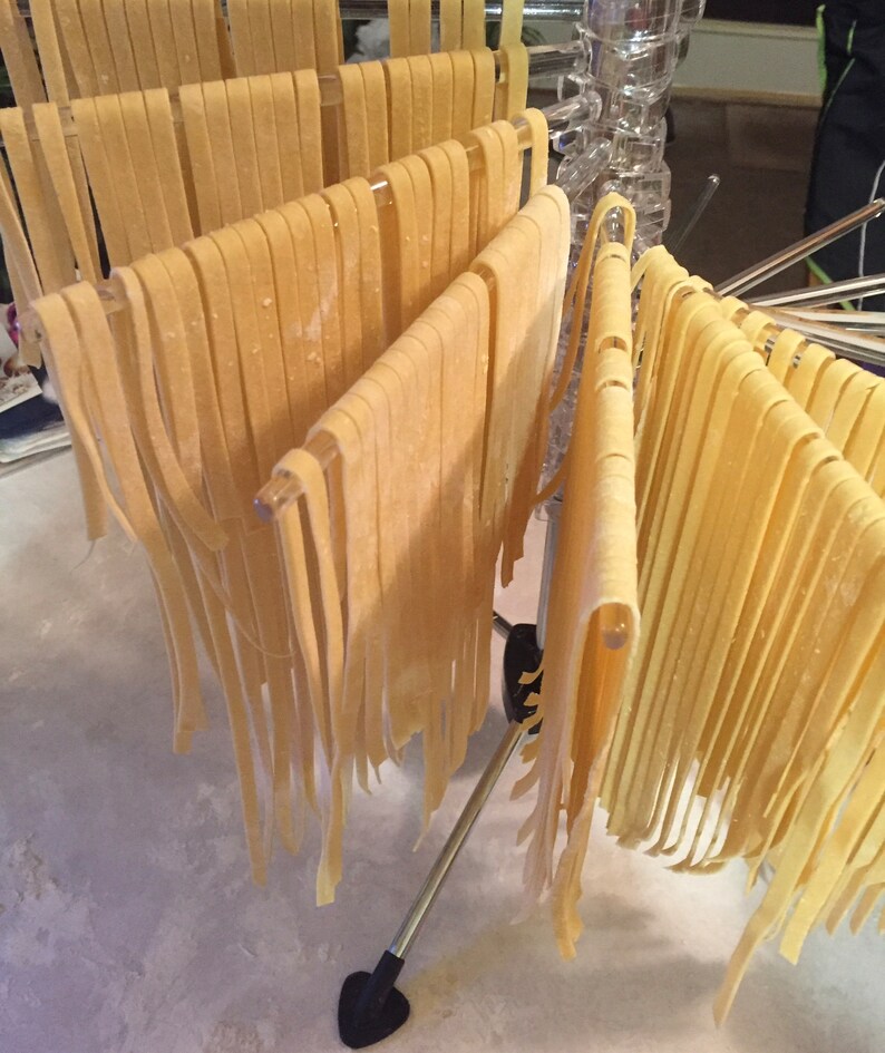 May include: Freshly made pasta noodles hanging on a drying rack. The noodles are a light yellow color and are arranged in neat rows. The rack is made of metal and plastic, and the background is a light-colored surface.