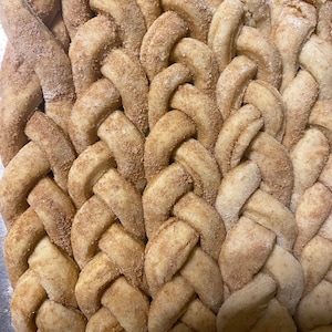 May include: Close-up of a braided bread loaf, dusted with cinnamon and sugar. The bread has a golden-brown color with a textured surface, showcasing a detailed weave pattern. The baked goods are a delicious treat.