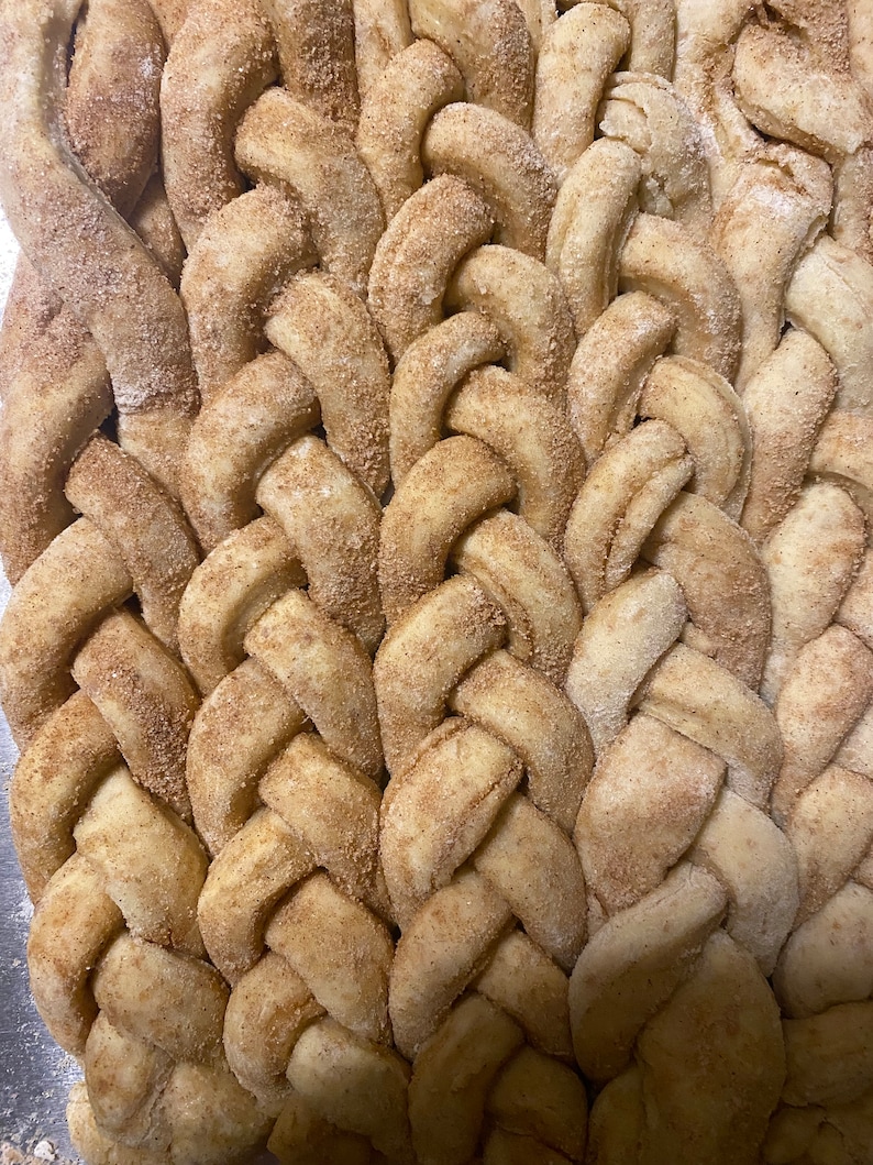 May include: Close-up of a braided bread loaf, dusted with cinnamon and sugar. The bread has a golden-brown color with a textured surface, showcasing a detailed weave pattern. The baked goods are a delicious treat.