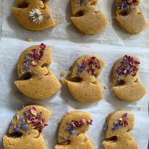 May include: Nine light brown, abstract-shaped pastries, each decorated with dried rose petals, blue cornflowers, and a single daisy. The pastries are arranged on a sheet of white parchment paper, creating a visually appealing arrangement.