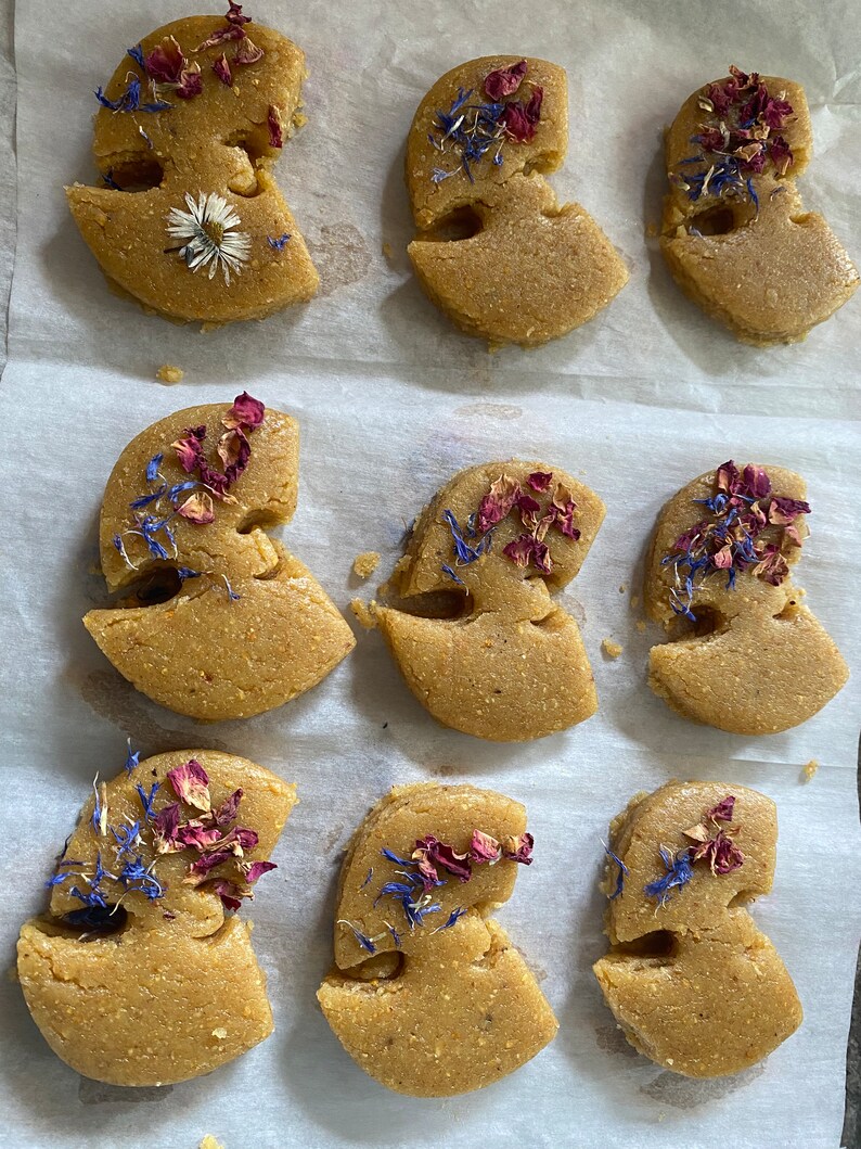 May include: Nine light brown, abstract-shaped pastries, each decorated with dried rose petals, blue cornflowers, and a single daisy. The pastries are arranged on a sheet of white parchment paper, creating a visually appealing arrangement.