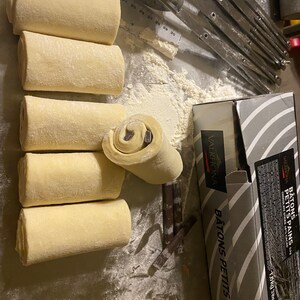 May include: Several unbaked pastries with chocolate filling are arranged on a metal surface dusted with flour. A partially cut pastry reveals the chocolate inside. Baking tools and a box of chocolate sticks are also visible.