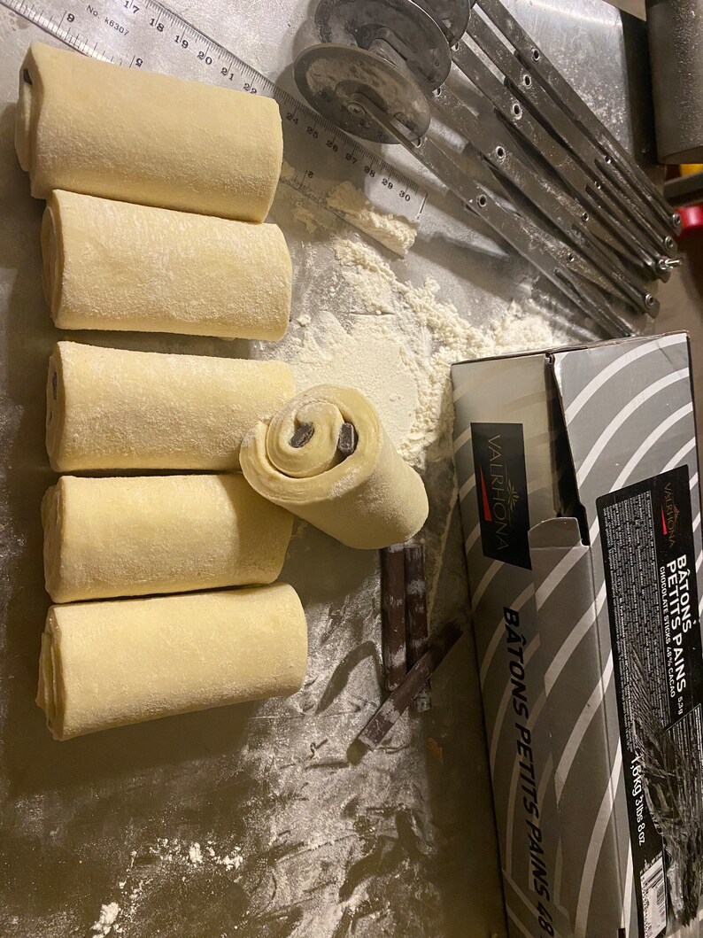 May include: Several unbaked pastries with chocolate filling are arranged on a metal surface dusted with flour. A partially cut pastry reveals the chocolate inside. Baking tools and a box of chocolate sticks are also visible.