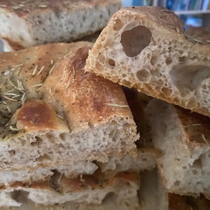May include: Close-up of freshly baked focaccia bread, cut into rectangular slices. The bread has a golden-brown crust and is topped with rosemary. The interior shows a porous texture. A delicious, homemade food item.