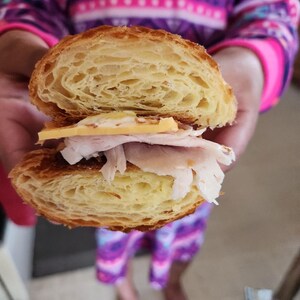 May include: A croissant sandwich cut in half, filled with sliced turkey, cheese, and a spread. The flaky, golden-brown croissant is layered with the sandwich fillings. The sandwich is held in front of a person.