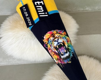 May include: A yellow and navy blue school cone with a colorful lion graphic. The cone has a blue trim and a tag that says "Emil" and another tag with the date "17.09.2026". The top is tied with a blue ribbon.