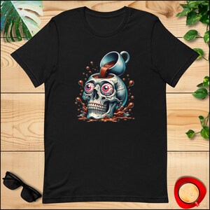 Op de afbeelding: Een zwart T-shirt met een afbeelding van een schedel met koffie die in zijn oogkassen wordt gegoten. De schedel heeft een verraste uitdrukking en de koffie stroomt uit de kop.