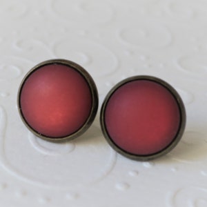 Könnte beinhalten: Ein Paar runde Ohrstecker. Jeder Ohrring hat einen matten roten Mittelteil, der in einen bronzefarbenen Metallrahmen gefasst ist. Die Ohrringe werden auf einer weißen, strukturierten Oberfläche präsentiert.