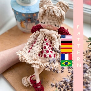 Könnte beinhalten: Häkelpuppenmuster in mehreren Sprachen. Die Puppe trägt ein beiges Kleid mit roten Punkten und einen roten Häkelumhang. Die Puppe hat blonde Haare und geschlossene Augen. Das Muster ist in Englisch, Deutsch, Spanisch, Französisch und Portugiesisch erhältlich.