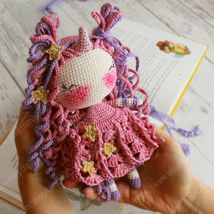 Puede incluir: Una mu&ntilde;eca de unicornio de crochet con pelo de lana rosa y morado, un vestido rosa con detalles de estrellas doradas y un cuerno blanco.
