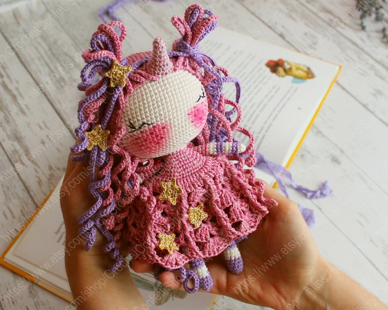 Puede incluir: Una mu&ntilde;eca de unicornio de crochet con pelo de lana rosa y morado, un vestido rosa con detalles de estrellas doradas y un cuerno blanco.