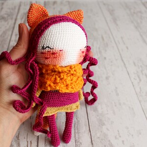 May include: A handmade crochet doll with a white face, pink cheeks, and closed eyes. The doll has curly pink hair, an orange scarf, and a pink and gold dress. Orange ears are attached to the top of the head. The doll is held in a hand.