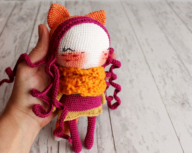 May include: A handmade crochet doll with a white face, pink cheeks, and closed eyes. The doll has curly pink hair, an orange scarf, and a pink and gold dress. Orange ears are attached to the top of the head. The doll is held in a hand.