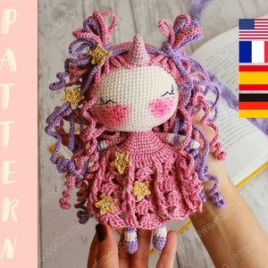 Puede incluir: Una mu&ntilde;eca de unicornio de crochet con pelo rosa y morado, un vestido rosa con estrellas doradas y una cara blanca. La mu&ntilde;eca est&aacute; siendo sostenida por una mano.