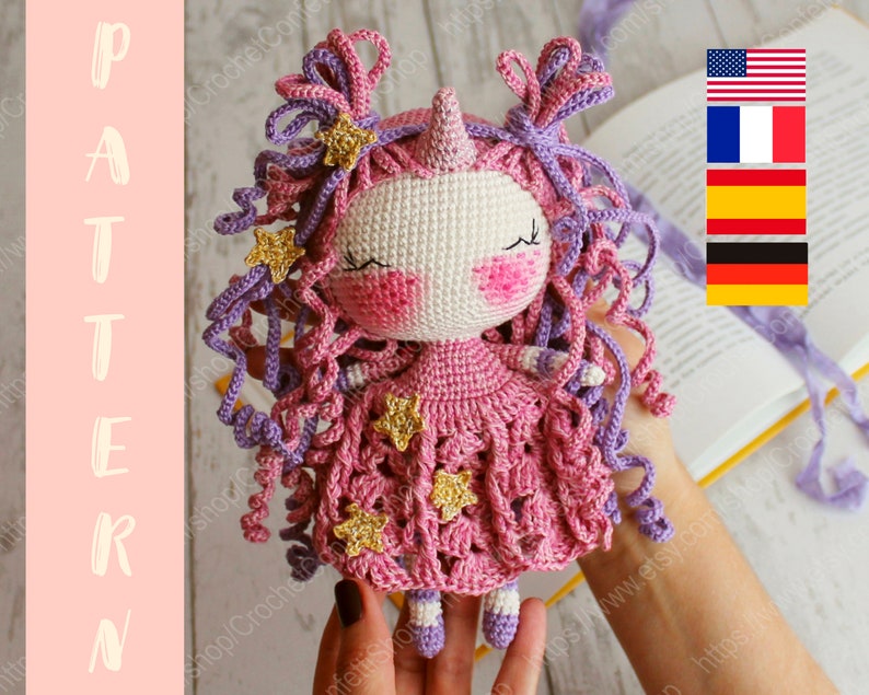 Puede incluir: Una mu&ntilde;eca de unicornio de crochet con pelo rosa y morado, un vestido rosa con estrellas doradas y una cara blanca. La mu&ntilde;eca est&aacute; siendo sostenida por una mano.