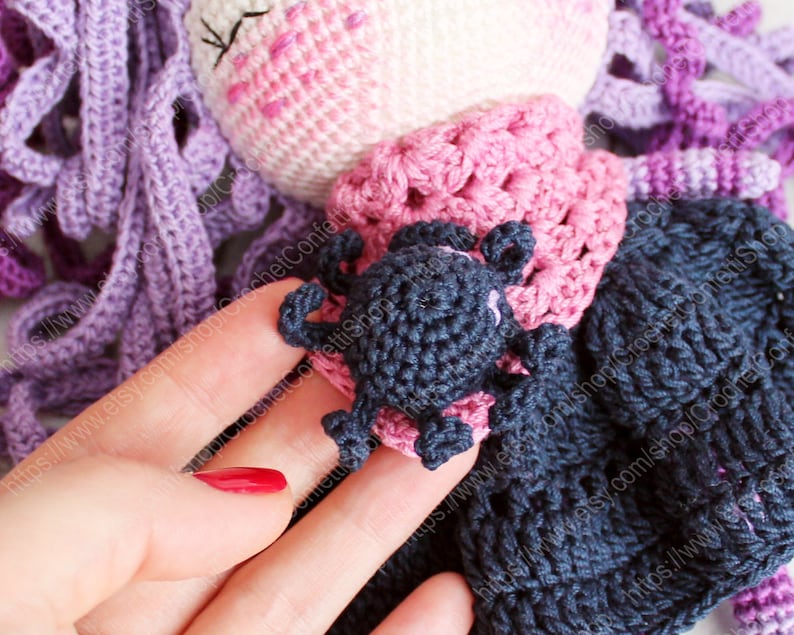 K&ouml;nnte beinhalten: Eine geh&auml;kelte Puppe mit lila Haaren, einem rosa Kleid und einer schwarzen geh&auml;kelten Spinne auf der Schulter.