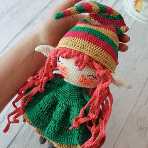 Peut inclure: Une poup&eacute;e elfe en crochet avec des cheveux roux, une robe verte et un chapeau ray&eacute;. La poup&eacute;e tient une &eacute;charpe rouge dans ses mains.