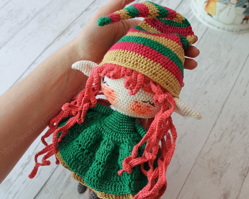 Peut inclure: Une poup&eacute;e elfe en crochet avec des cheveux roux, une robe verte et un chapeau ray&eacute;. La poup&eacute;e tient une &eacute;charpe rouge dans ses mains.