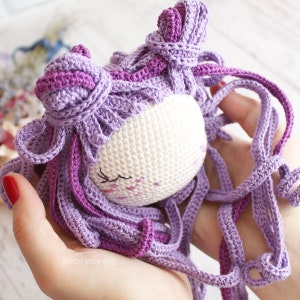 K&ouml;nnte beinhalten: Eine geh&auml;kelte Puppe mit lila Haaren und einem wei&szlig;en Gesicht. Die Puppe hat geschlossene Augen und eine kleine rosa Nase. Die Haare sind lang und flie&szlig;end, mit zwei lila Schleifen auf dem Kopf.