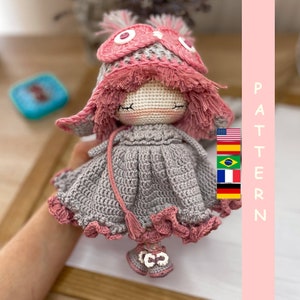 Pode incluir: Boneca de crochê com roupa rosa e cinza. A boneca está usando um chapéu rosa com franjas e um vestido cinza com acabamento rosa. A boneca tem uma cauda de fio cinza.