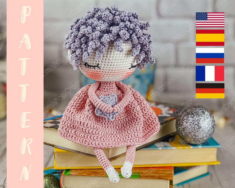 May include: Crocheted doll with purple hair, wearing a pink dress and holding a purple heart. The doll is sitting on a stack of books.
