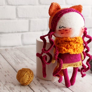 May include: A handmade crochet doll with a white face, rosy cheeks, and closed eyes. The doll has orange cat ears, curly magenta hair, and a yellow and orange scarf. It is wearing a magenta dress with a yellow trim.