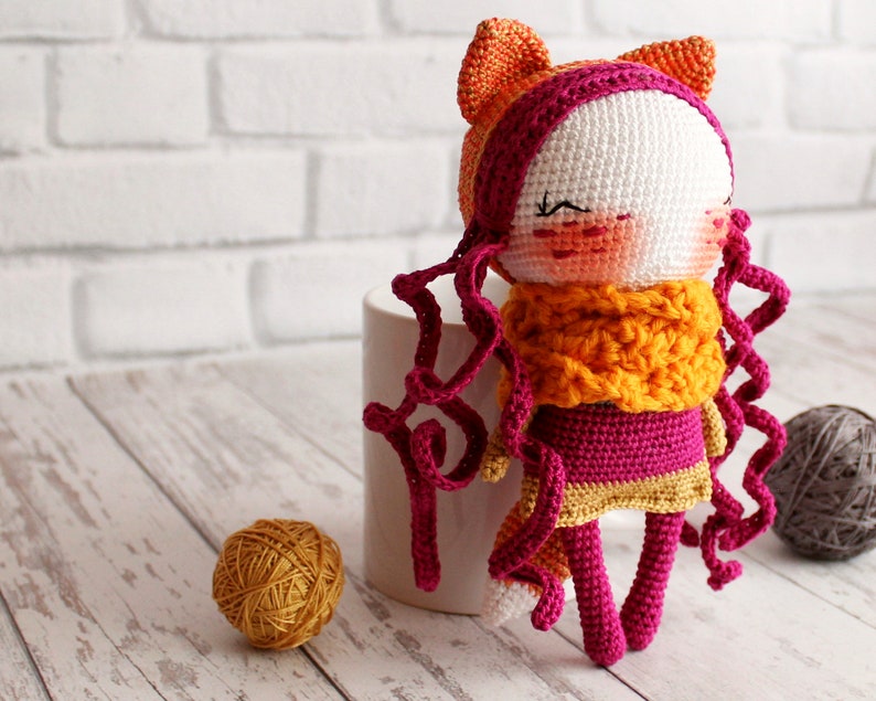 May include: A handmade crochet doll with a white face, rosy cheeks, and closed eyes. The doll has orange cat ears, curly magenta hair, and a yellow and orange scarf. It is wearing a magenta dress with a yellow trim.