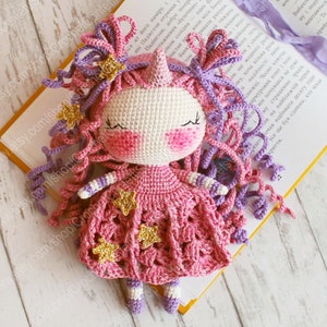 Puede incluir: Una mu&ntilde;eca de unicornio de crochet que lleva un vestido rosa con estrellas doradas. La mu&ntilde;eca tiene el pelo de lana morado y rosa y una cara blanca con mejillas rosas.