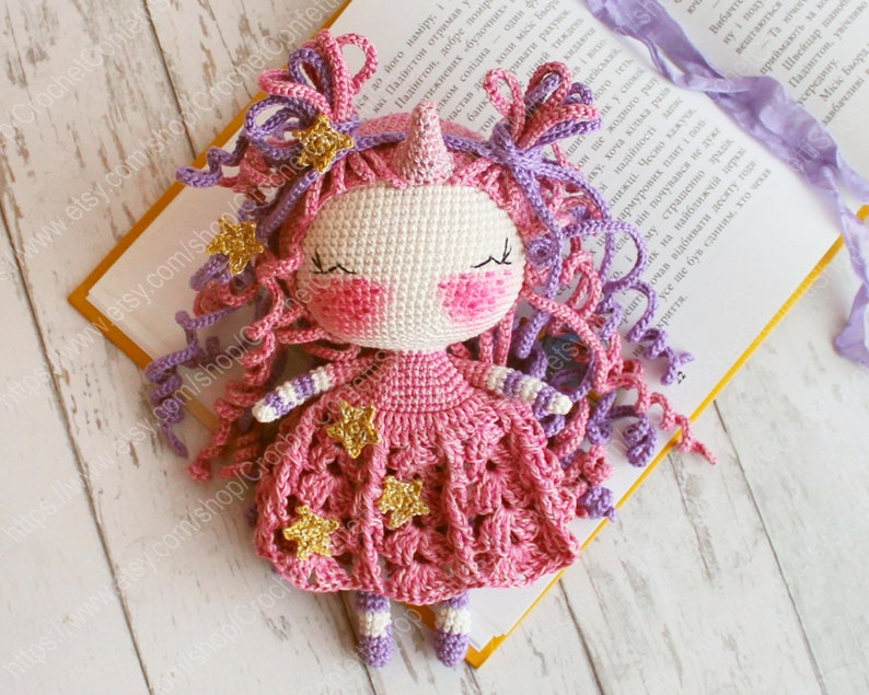 Puede incluir: Una mu&ntilde;eca de unicornio de crochet que lleva un vestido rosa con estrellas doradas. La mu&ntilde;eca tiene el pelo de lana morado y rosa y una cara blanca con mejillas rosas.