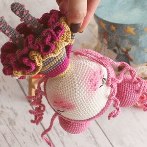Pu&ograve; includere: Un giocattolo di unicorno all'uncinetto con una criniera rosa e una corona rosa e gialla. L'unicorno &egrave; fatto con filato bianco, rosa e giallo.