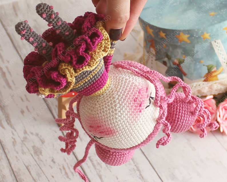 Pu&ograve; includere: Un giocattolo di unicorno all'uncinetto con una criniera rosa e una corona rosa e gialla. L'unicorno &egrave; fatto con filato bianco, rosa e giallo.