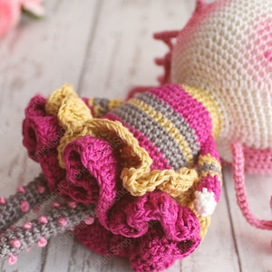 Pu&ograve; includere: Una bambola all'uncinetto con un vestito a righe rosa e grigio e una gonna a balze gialla. La bambola ha una testa bianca con guance rosa e un fiocco rosa all'uncinetto.