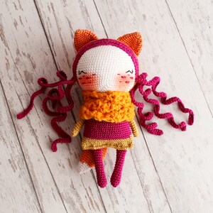 May include: A handmade crochet doll with a fox-like design. The doll features a white face, pink cheeks, and closed eyes. It wears a pink and gold dress, an orange scarf, and has orange ears. Long, curly pink yarn strands serve as hair.