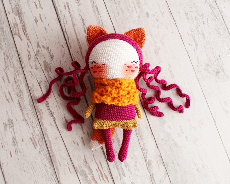 May include: A handmade crochet doll with a fox-like design. The doll features a white face, pink cheeks, and closed eyes. It wears a pink and gold dress, an orange scarf, and has orange ears. Long, curly pink yarn strands serve as hair.