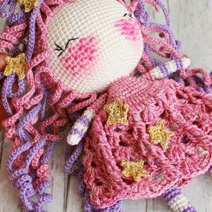 Puede incluir: Una mu&ntilde;eca de crochet con pelo rosa y morado, que lleva un vestido rosa con detalles de estrellas doradas.
