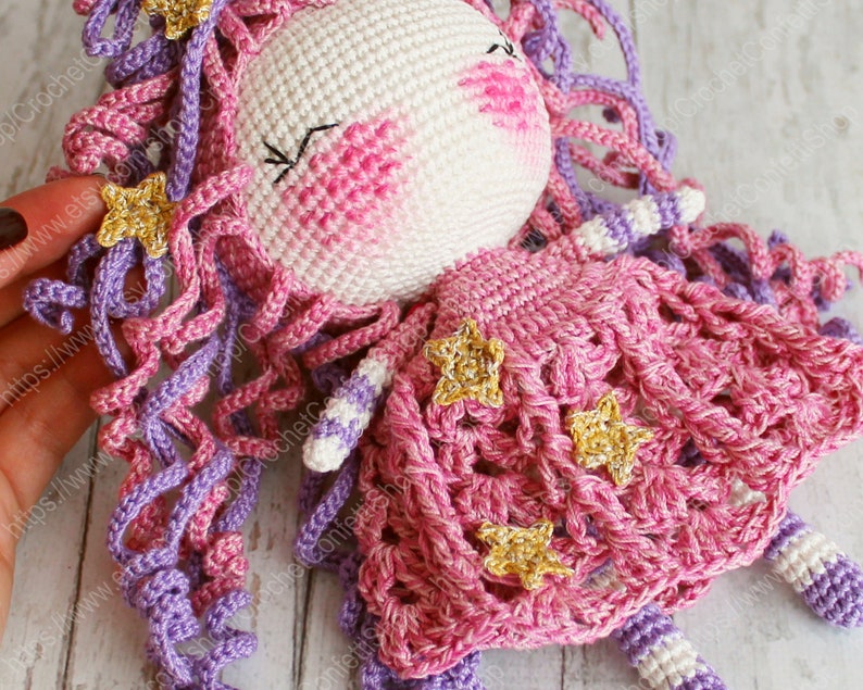 Puede incluir: Una mu&ntilde;eca de crochet con pelo rosa y morado, que lleva un vestido rosa con detalles de estrellas doradas.
