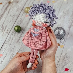 May include: A crocheted doll with purple curly hair, wearing a pink dress and holding a heart-shaped object. The doll has a white face with closed eyes and rosy cheeks.