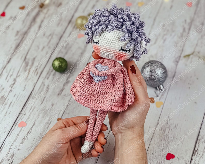 May include: A crocheted doll with purple curly hair, wearing a pink dress and holding a heart-shaped object. The doll has a white face with closed eyes and rosy cheeks.