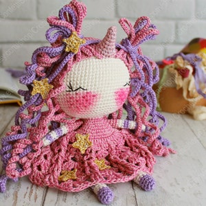 Puede incluir: Una mu&ntilde;eca de unicornio de crochet con pelo rosa y morado, un vestido rosa con estrellas doradas y un cuerno blanco. La mu&ntilde;eca est&aacute; sentada sobre una superficie de madera.