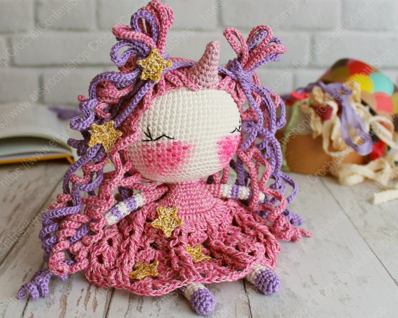 Puede incluir: Una mu&ntilde;eca de unicornio de crochet con pelo rosa y morado, un vestido rosa con estrellas doradas y un cuerno blanco. La mu&ntilde;eca est&aacute; sentada sobre una superficie de madera.
