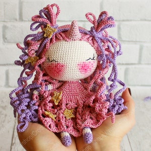Puede incluir: Una mu&ntilde;eca de unicornio de crochet con pelo rosa y morado, un vestido rosa con estrellas doradas y una cara blanca con los ojos cerrados.