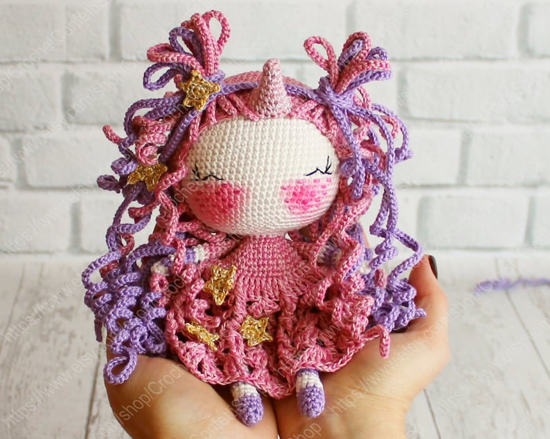 Puede incluir: Una mu&ntilde;eca de unicornio de crochet con pelo rosa y morado, un vestido rosa con estrellas doradas y una cara blanca con los ojos cerrados.