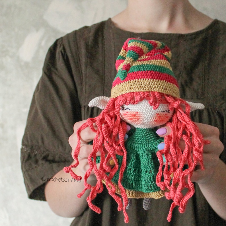 Peut inclure: Une poup&eacute;e elfe en crochet avec des cheveux roux, une robe verte et un chapeau ray&eacute;. La poup&eacute;e est tenue par les mains d'une personne.