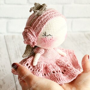 Op de afbeelding: Een gehaakte pop die een roze jurk en een kroon draagt. De pop heeft een wit gezicht met roze wangen en gesloten ogen.